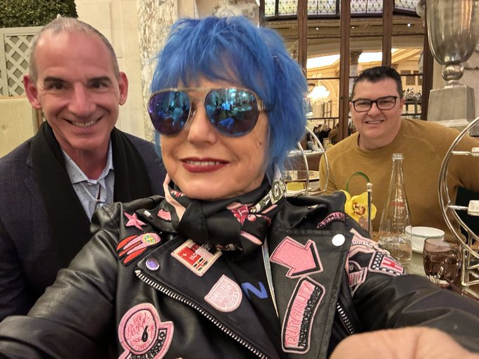 A selfie of a woman with bright blue hair and reflective sunglasses wearing a black leather jacket with pink patches, sitting between two smiling men at a restaurant.