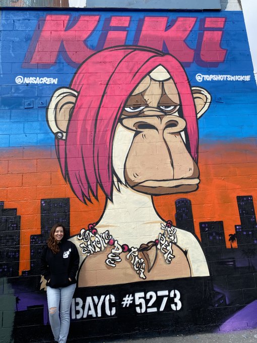 A woman stands smiling beside a vibrant street art mural featuring a pink-haired Bored Ape with the text "KIKI," "@NASACREW," "@TOPSHOTSWICKIE," and "BAYC #5273," directly referencing Bored Ape Yacht Club NFT #5273. The artwork, tagged "Bored by Kiki est 2021," showcases the artist's 2021 series celebrating the iconic BAYC collection.
