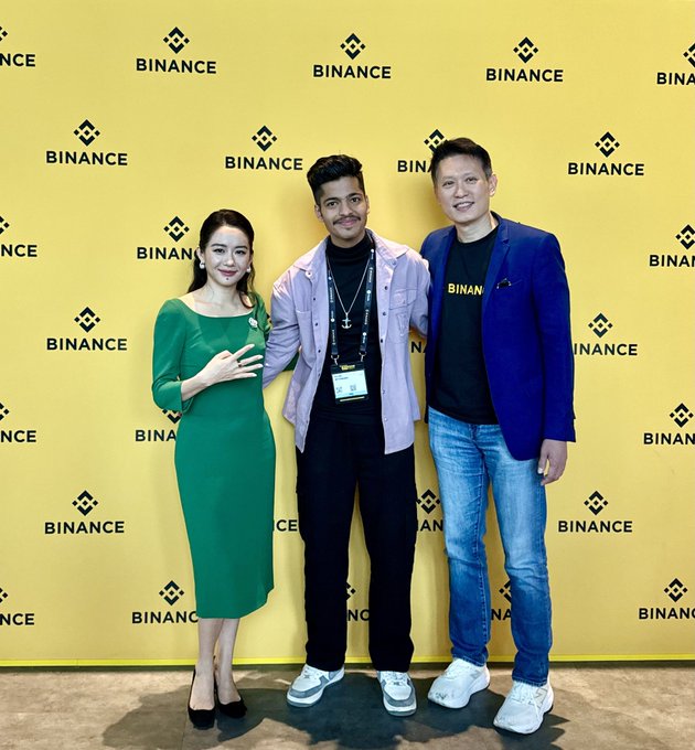 A photo of three people posing together in front of a yellow backdrop branded with the Binance logo, including a man in a 'BINAN' t-shirt and blazer.