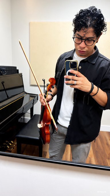 A young man with curly dark hair and glasses stands in a room with a piano, holding a violin and bow while taking a mirror selfie with his smartphone.