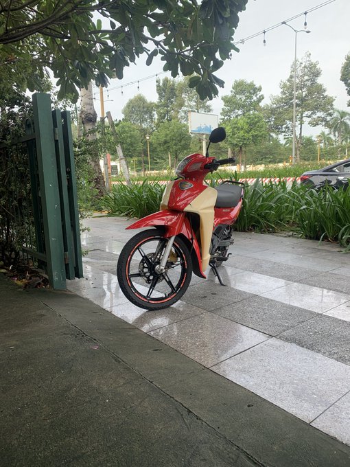 A red and gold motorbike is parked on a wet tiled sidewalk near a green gate, ready to take the rider out for a morning coffee ("Cafe sáng thôi cả nhà ơi" — "Just morning coffee, everyone").