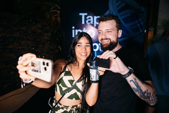 The image shows a man holding up his phone, taking a selfie with another person who is also holding their phone. The background of the photo features a billboard that says "TAP into something" and has an image of a plant on it. This suggests that they might be at an event or gathering where people are encouraged to take photos and share them on social media platforms, possibly promoting a product or service related to plants or nature.

The post caption reads: Should I try @Tangem? @matheus_slivak". The caption is likely intended to encourage the viewer to engage with the image by taking a photo of themselves and tagging it with the relevant hashtag.