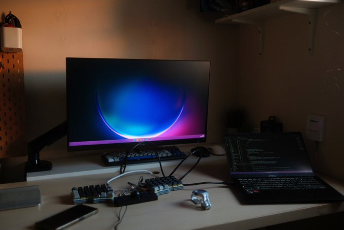 A dim home-office setup includes a large monitor displaying a colorful abstract wallpaper, a laptop running code, split mechanical keyboards, a mouse, and a smartphone.