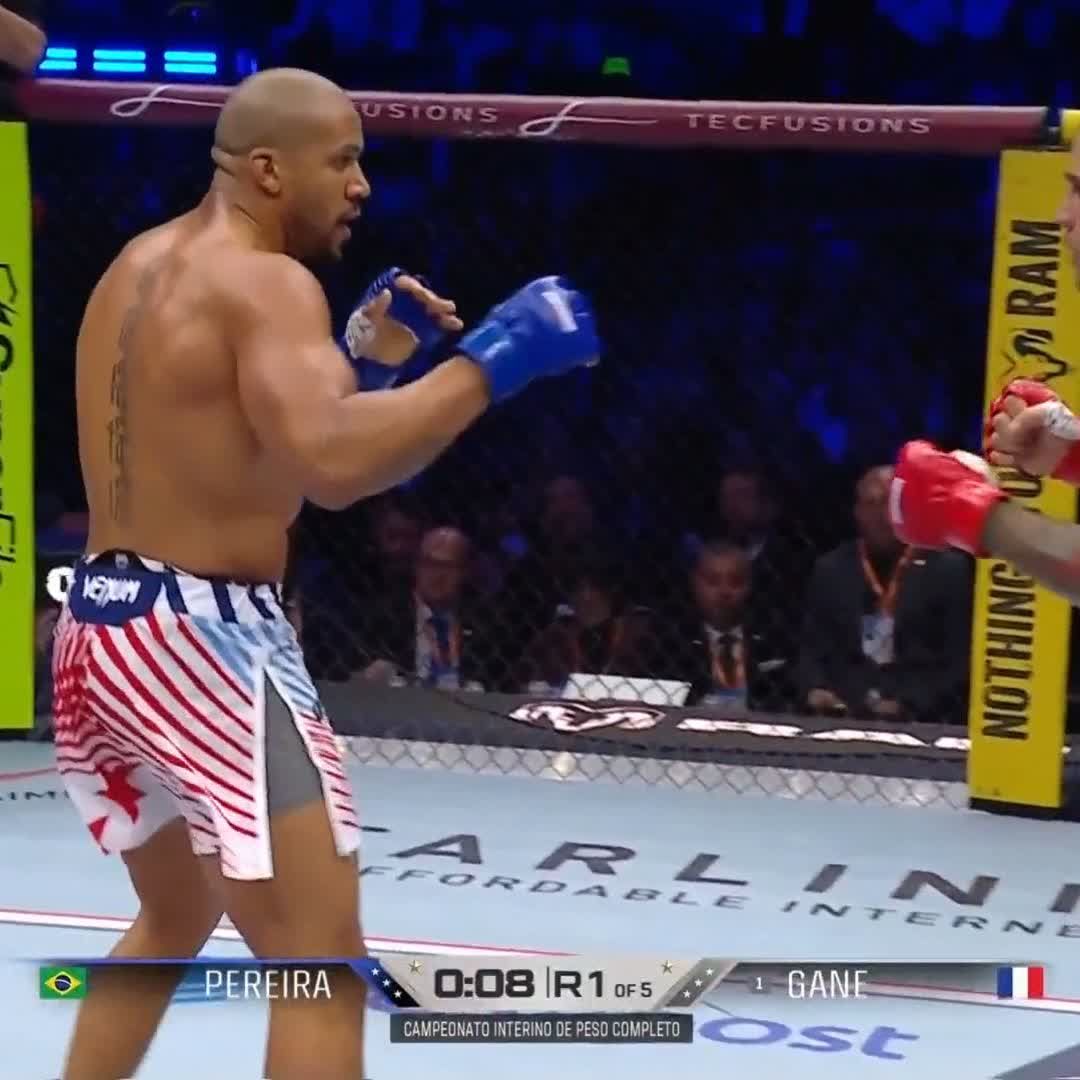 The image shows a man wearing red shorts, standing in the middle of a ring during a fight at a professional MMA event. He is holding a pair of boxing gloves and appears to be preparing for an upcoming punch. The fighter's stance suggests he is ready to engage with his opponent. In the background, there are several people watching the match, indicating that it might be a significant event or a popular sport.