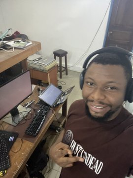 A smiling man wearing headphones and a brown "RASTIC DEAD" t-shirt sits at a desk with computer monitors and a keyboard, posing for a selfie.