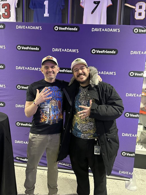 Two men posing at what appears to be a Dave & Adam's / VeeFriends event, with one wearing a cap and a graphic t-shirt, and the other wearing a fur-hooded jacket, with sports jerseys visible on the wall behind them.