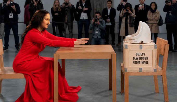 A woman in a long red dress kneels at a wooden table with her hands resting on it, while a white toilet sits atop a wooden crate labeled "OBJECT MAY DIFFER FROM IMAGE" with a crowd of photographers in the background.
