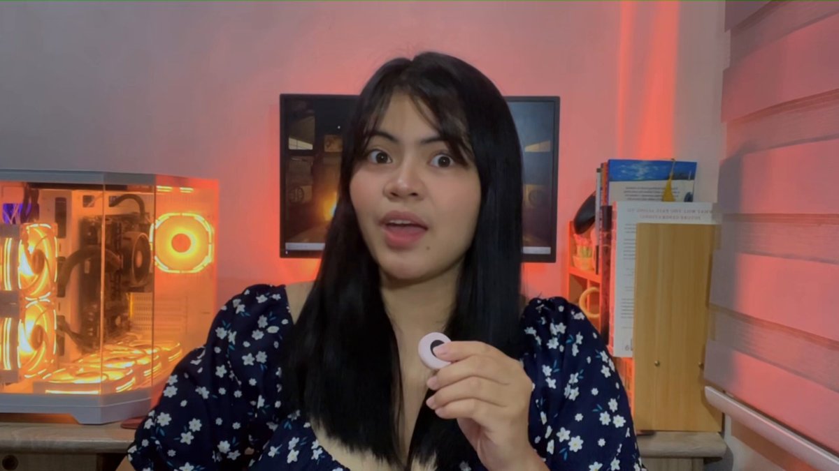 The image features a woman wearing a blue floral top holding up a small white object with her right hand while standing in front of a computer screen displaying a video. The background includes a desk, bookshelf, and a window, suggesting that she might be working or studying from home. The post about the Injective project is displayed on the image's caption, which reads: "my first Injective video I broke it down in simple words in this video. It is a blockchain built for trading and finance, aiming to make trading faster, more open, and fully on-chain".