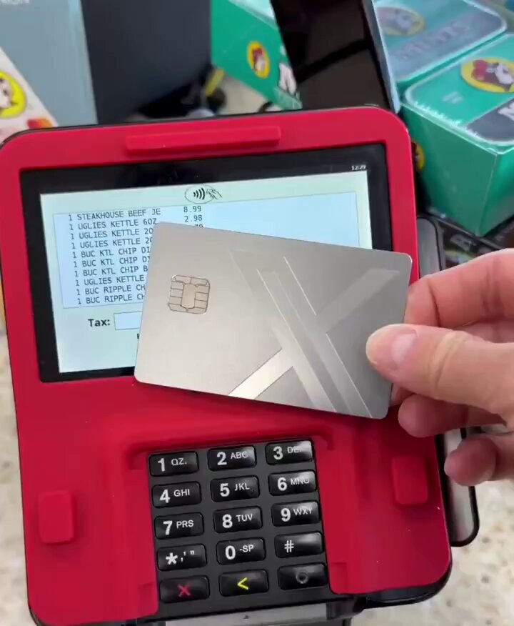 The image shows a person holding a silver credit card, which appears to be a Mastercard, over a red cash register. The credit card has been used for making a purchase at a store or market. The text in the image suggests that this is part of a new payment system called Bitcoin Money, which could potentially replace traditional currency and change the way people make transactions. This development might lead to real-world adoption of Bitcoin Money as a form of payment, offering users greater financial flexibility and security.