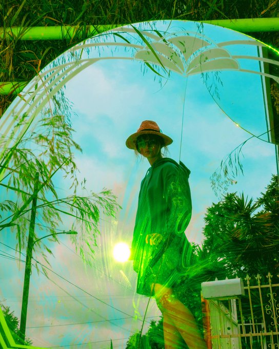 A woman wearing a hat and dark coat stands under a decorative green metal archway with leaf motifs, captured with a green-tinted lens flare effect.