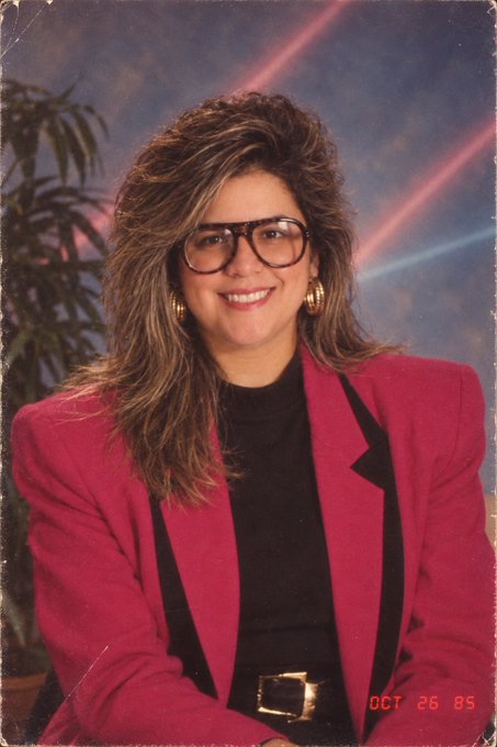 A vintage 1980s portrait dated 'OCT 26 85' showing a smiling woman with voluminous highlighted hair, large eyeglasses, gold earrings, and a bright pink blazer with black trim over a black top.