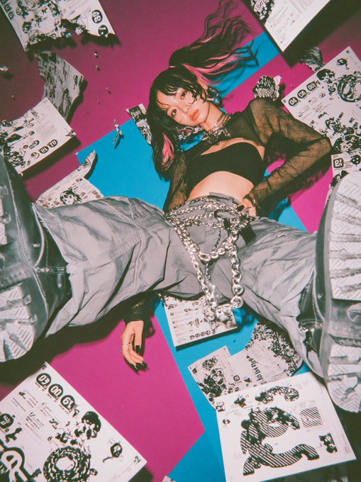 A young woman with dark hair featuring pink highlights lies on a vibrant pink and blue background, surrounded by scattered papers, flyers, and "Turnstile" tickets. She wears a black mesh top, silver chain belt, and oversized gray boots, striking a bold pose that matches the invite-like caption "Come on over."