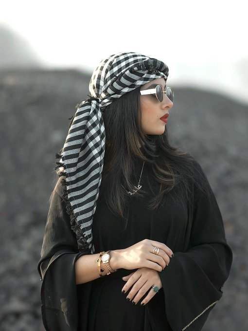 A stylish woman wearing a black and white checkered keffiyeh-style headscarf, a black flowing garment, and white-framed round sunglasses, standing against a dark rocky background.