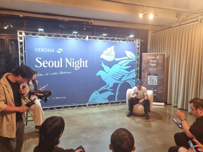 An indoor event space features a large blue 'Verona Seoul Night' banner with a QR code, where attendees watch as a person sits on a white chair being filmed by a videographer.