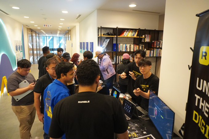 A group of attendees gathers around a technology booth, with visible text including "RAREIBLE" on a shirt and partial words "UNI," "THE," and "DIGI" on a banner, capturing the networking atmosphere of the event organized by SuperteamMY, Solana Spaces, and Superscrypt.