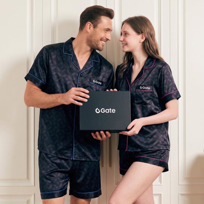 A man and woman in matching dark patterned silk pajama sets with blue and pink piping, respectively, both featuring "Gate" logos, jointly holding a black box with the "Gate" logo.