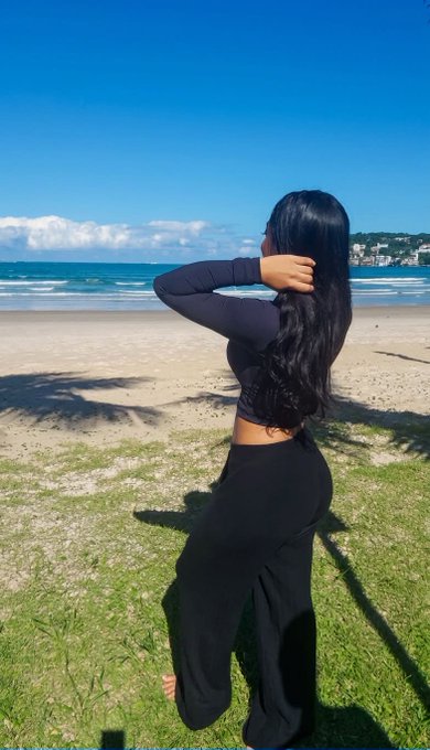 A woman with long dark hair stands facing away on a grassy area overlooking a sandy beach and the ocean under a clear blue sky.
