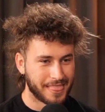 A young man with curly brown hair and a beard smiles while wearing a black shirt and an earring, appearing to be in a studio setting.