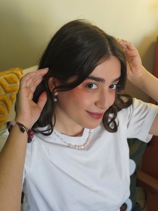 A young woman with long dark hair wearing a white t-shirt and a pearl necklace, adjusting her hair while looking at the camera, with a bracelet visible on her wrist.