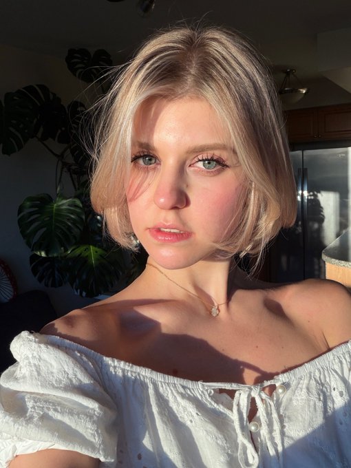 A selfie of a young woman with blonde hair and green eyes wearing a white off-shoulder top and a delicate necklace, with a monstera plant visible in the background.