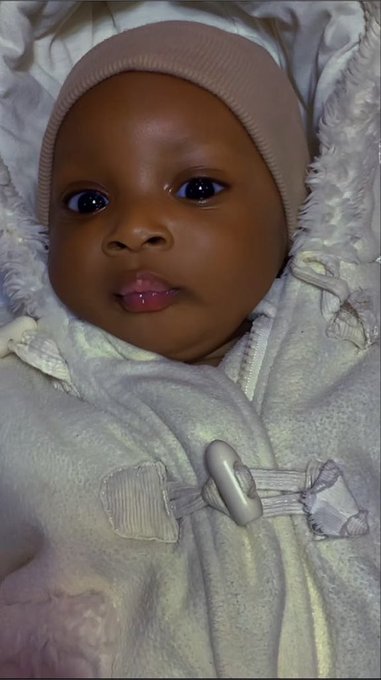 A close-up portrait of a baby with dark skin wearing a pink ribbed beanie hat and wrapped in a cream-colored plush blanket with a decorative bow clasp.