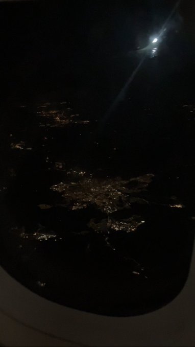 An aerial nighttime photograph taken from an airplane window showing city lights below and part of the plane's wing illuminated against the dark sky.