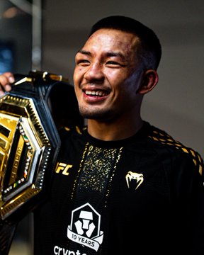 Joshua Van smiles broadly, wearing a black Crypto.com sponsored jersey with "NO YEARS" and "crypto" text visible, while holding a UFC championship belt featuring gold and silver plates with his name "VAN" displayed on it.