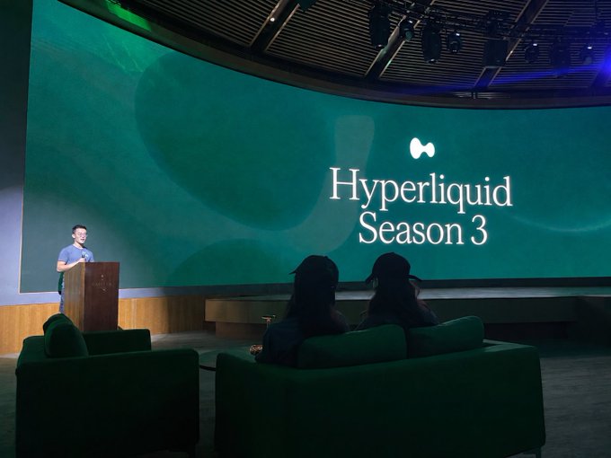 A presentation event for 'Hyperliquid Season 3' displayed on a large green screen, with a speaker at a podium addressing an audience seated on green sofas.
