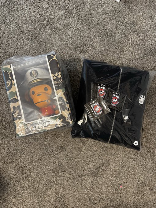 Two pieces of BAPE streetwear merchandise in clear plastic packaging on a gray carpet: a camo zip-up hoodie featuring the Baby Milo character in a captain's outfit, and a black jacket.
