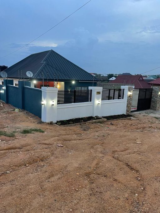 A modern single-story bungalow house with a dark gray hipped roof, white walls, black metal fence, and illuminated pillars, located in a neighborhood surrounded by unpaved dirt roads under a cloudy sky.