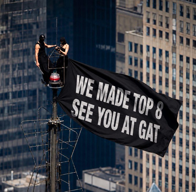 Two people in black outfits perch atop a tall structure, unfurling a large black banner that reads "WE MADE TOP 8 SEE YOU AT GAT" against a city skyline backdrop, celebrating their achievement of advancing to the Top 8 in the Crypto Stage and securing their spot at the GAT competition.