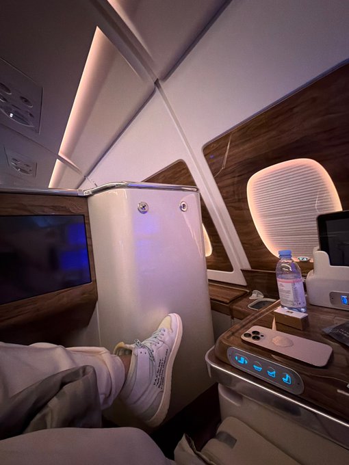 A passenger relaxes in what appears to be a luxurious first-class or business-class cabin, with their white sneaker propped up on a footrest, a water bottle, and a phone on a polished wooden side console featuring illuminated seat controls, while the post humorously contrasts this lavish travel experience with a risky 120k memecoin pump-and-dump gamble.