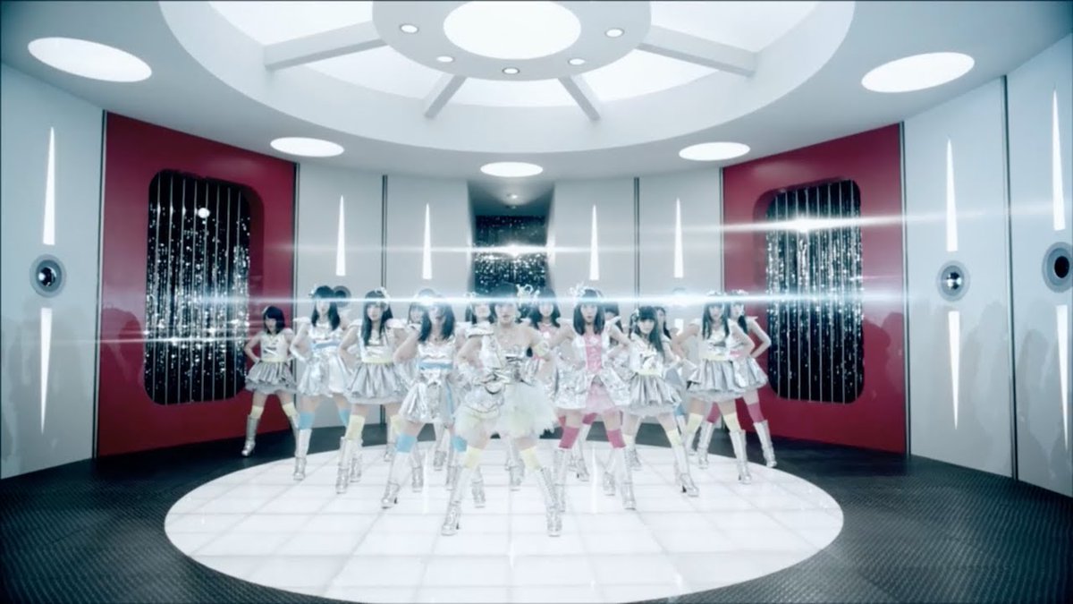 A group of female performers in shiny silver costumes performing in a futuristic white circular room, likely a J-pop music video scene.