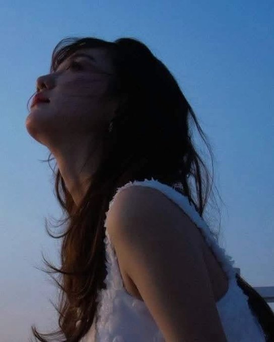 A young woman with long dark hair in a white top looks up against a clear blue sky during golden hour.