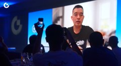 A low-resolution image of a man speaking at a conference or presentation with an audience visible in the foreground.