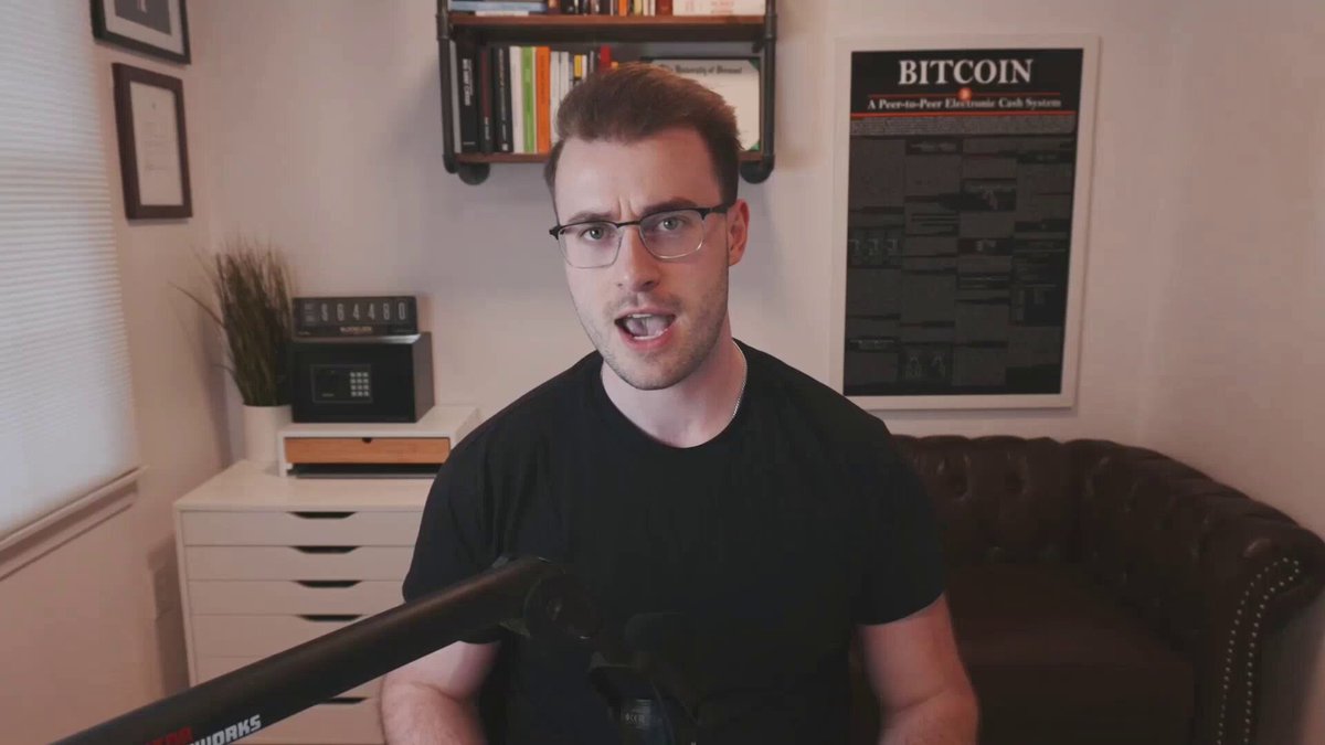 In this image, a man wearing glasses and a black shirt is standing next to a couch, holding a microphone. The man appears to be talking or recording something with the microphone. Behind him, there are bookshelves filled with various books arranged on them. Another person can also be seen sitting in a chair at the back of the room.