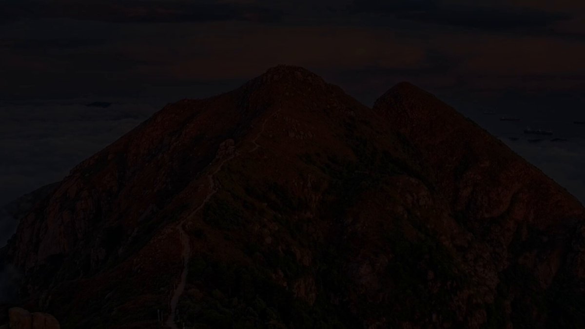 The image shows a mountain range with two peaks, one of which has a trail leading up to it. The mountains are dark brown, suggesting they could be granite or similar types of rock. The sky above them appears to be cloudy, indicating an overcast day. The image is taken from a low angle, making the mountains appear larger and more imposing.