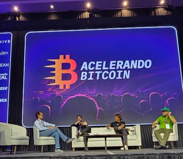 A panel discussion on stage at the 'Acelerando Bitcoin' conference, with four speakers seated in chairs in front of a large Bitcoin-themed backdrop.
