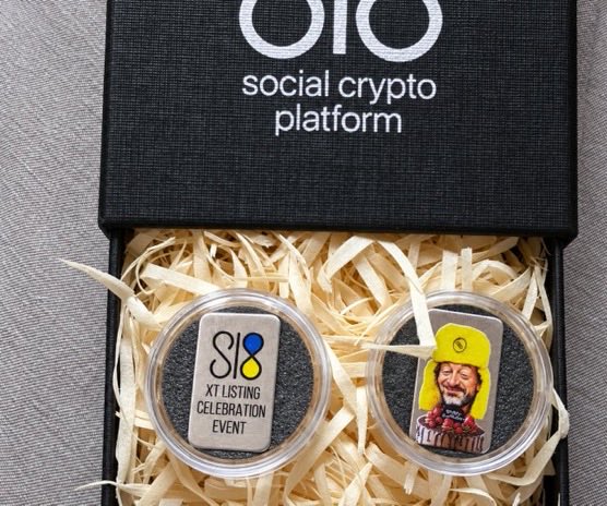 An open presentation box from 'oro social crypto platform' containing two commemorative coins in protective capsules, one labeled 'XT LISTING CELEBRATION EVENT' and the other featuring a cartoon figure wearing a yellow cap.