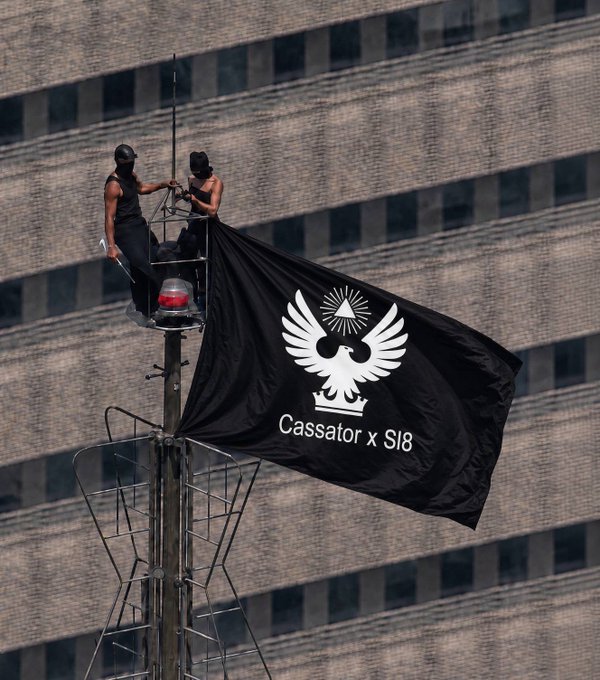 Two individuals in black attire perch atop a streetlight fixture, unfurling a large black flag featuring a white eagle emblem and the text "Cassator x Sl8," with a tall building facade in the background. The image visually represents the Sl8 Social brand mentioned in the post, showcasing a bold promotional display for the Stellar-based social platform.