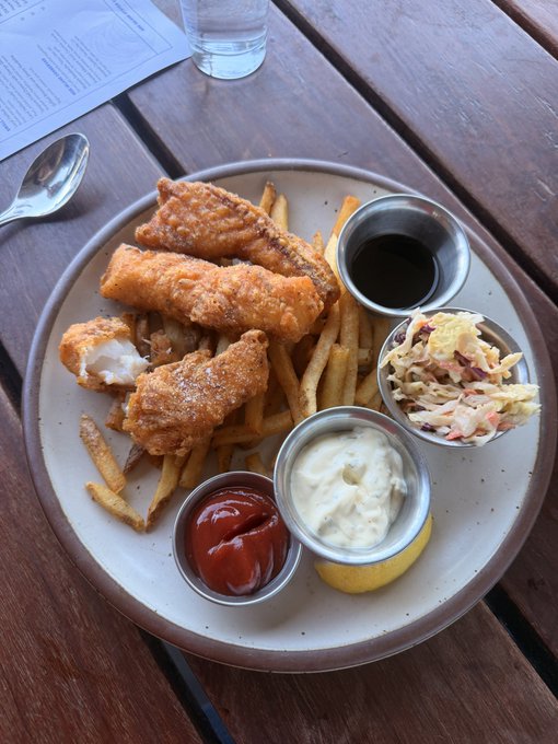 A plate of fish and chips with coleslaw, ketchup, mayonnaise, tartar sauce, and a lemon wedge sits on a wooden table beside a glass of water and silverware.