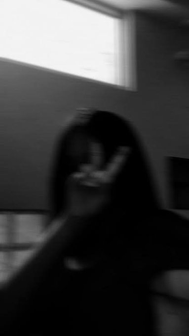 A blurry, black-and-white photo of a person wearing glasses and a hoodie making a peace sign with their hand.