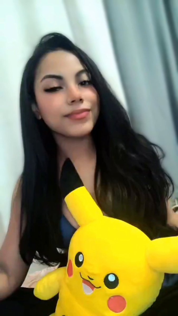 The image features a young woman with long hair holding a large yellow Pokemon plushie. She appears to be posing for a photo or video, possibly sharing her excitement about the Pokemon franchise. The background of the image shows a window, which could suggest that she is indoors in a room.