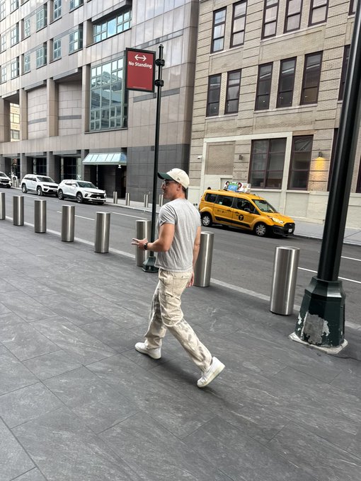 A man wearing a cap, grey t-shirt, and beige pants walks on a city sidewalk near a 'No Standing' sign, with a yellow NYC taxi and tall buildings in the background.