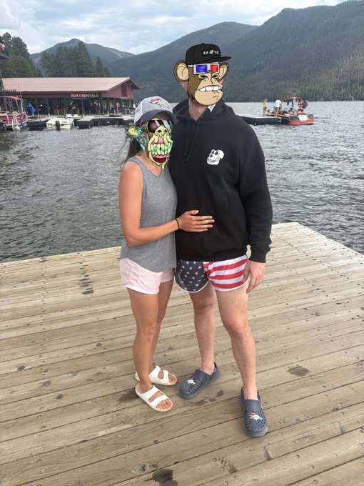 Two people stand on a wooden dock by a lake with mountains in the background; their faces are obscured by cartoon stickers, and one wears shorts with an American flag pattern.