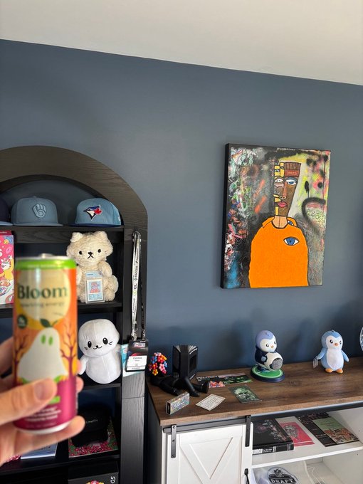 A hand holds an orange can labeled 'Bloom' in a room featuring a Basquiat-style painting of an orange torso on a blue wall, alongside shelves displaying baseball caps, a teddy bear, and plush toys.