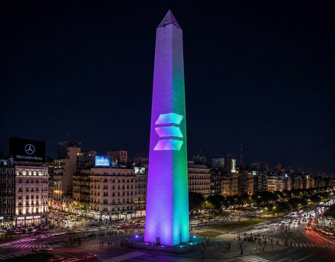 The Obelisco de Buenos Aires (Buenos Aires Obelisk) illuminated at night with purple, pink, and green lights displaying the Solana (SOL) cryptocurrency logo on its face.
