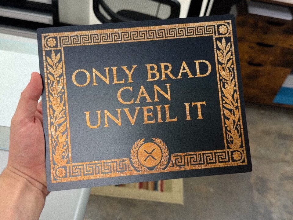 The image features a person holding up a black sign with gold lettering that reads "Only Brad Can Unveil It". The sign has an ornate design, including a shield and a laurel wreath at the bottom center of the frame. The background is blurred, but it appears to be a room or office setting.