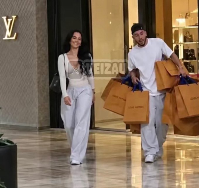 A man carries multiple Louis Vuitton shopping bags while walking alongside a woman leaving what appears to be a luxury store, exemplifying the extravagant shopping haul aesthetic that the "rasmr" trend—known for satisfying, sensory shopping unboxing videos—popularizes for content creators to imitate.