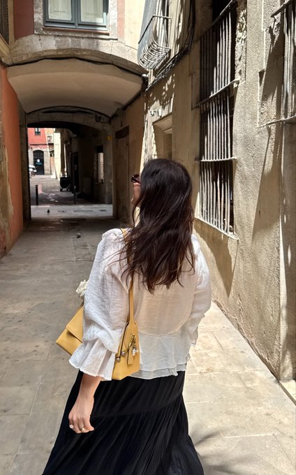 A woman with long dark hair wearing a white blouse and black skirt walks down a narrow stone alleyway carrying a yellow handbag, with arched passageways visible ahead.